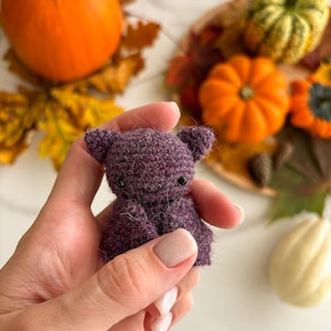 Op de afbeelding: Een kleine, donkerpaarse gehaakte vleermuisfiguur met een pluizige textuur, puntige oren en twee kleine zwarte kraaloogjes. Het handgemaakte item heeft een afgerond hoofd en gevouwen vleugels, met gedetailleerd haakwerk over de gehele vorm.