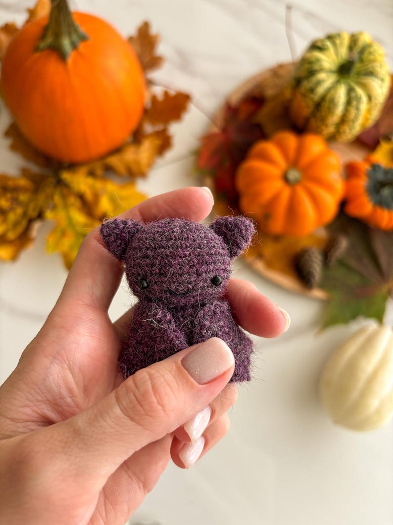 Op de afbeelding: Een kleine, donkerpaarse gehaakte vleermuisfiguur met een pluizige textuur, puntige oren en twee kleine zwarte kraaloogjes. Het handgemaakte item heeft een afgerond hoofd en gevouwen vleugels, met gedetailleerd haakwerk over de gehele vorm.