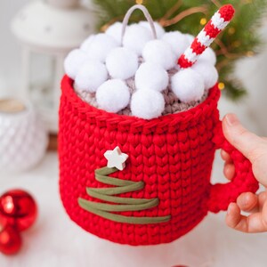 Pu&ograve; includere: Una tazza rossa all'uncinetto con un coperchio bianco all'uncinetto e un design di albero di Natale verde all'uncinetto. La tazza &egrave; piena di pompon bianchi e una cannuccia a righe rosse e bianche all'uncinetto.