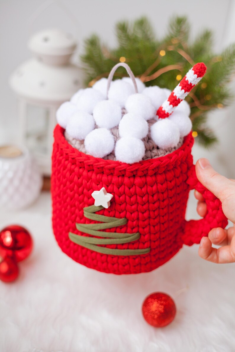Pu&ograve; includere: Una tazza rossa all'uncinetto con un coperchio bianco all'uncinetto e un design di albero di Natale verde all'uncinetto. La tazza &egrave; piena di pompon bianchi e una cannuccia a righe rosse e bianche all'uncinetto.