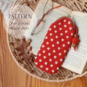Peut inclure: Étui à lunettes rouge crocheté à pois blancs, avec fermoir en métal et pompon de perles. Le texte "PATTERN *not a ready made item" est visible. L'étui repose sur un livre dans un panier tressé.