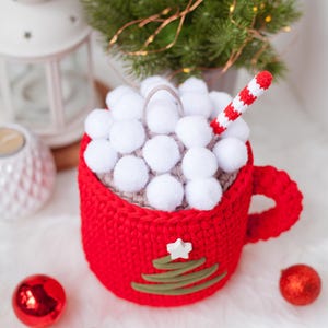 Pu&ograve; includere: Una tazza rossa all'uncinetto con un top a pompon bianco e un design a forma di albero di Natale verde all'uncinetto. La tazza ha una cannuccia a righe bianche ed &egrave; piena di pompon bianchi.