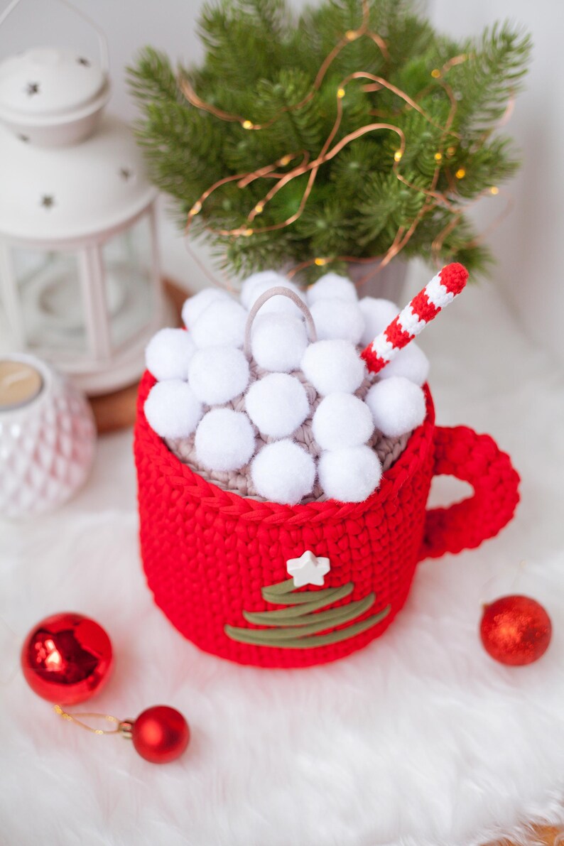 Pu&ograve; includere: Una tazza rossa all'uncinetto con un top a pompon bianco e un design a forma di albero di Natale verde all'uncinetto. La tazza ha una cannuccia a righe bianche ed &egrave; piena di pompon bianchi.