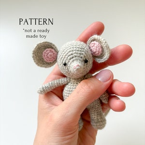 以下が含まれることがあります： ピンクの耳と黒い目をしたグレーの編みぐるみマウス。マウスは手で持たれています。画像には「PATTERN *not a ready made toy」というテキストが表示されています。