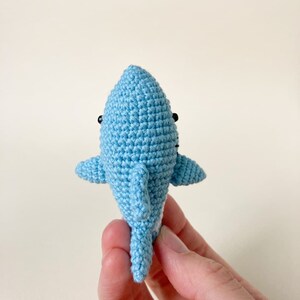 May include: A small, crocheted blue shark toy with black eyes. The shark is being held by a hand.