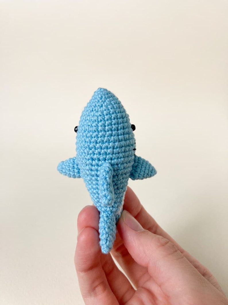 May include: A small, crocheted blue shark toy with black eyes. The shark is being held by a hand.