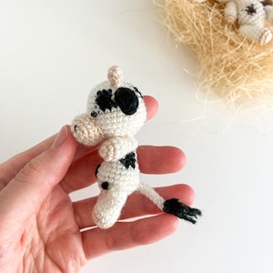 May include: A small crocheted cow toy with black and white spots. The cow is standing on its hind legs with its front legs raised. The cow has a black tail and a white face with black eyes and a black nose.