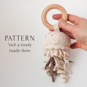 Op de afbeelding: Gehaakt kwallen speelgoed met een houten ring eraan. Het speelgoed is beige met bruine accenten. De tekst "PATTERN *not a ready made item" is zichtbaar in de afbeelding.