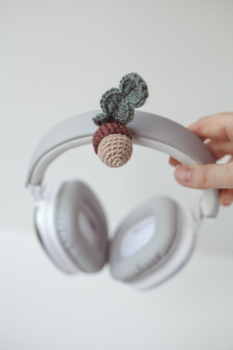 May include: White headphones with a crocheted acorn charm attached to the headband. The acorn is brown and green.