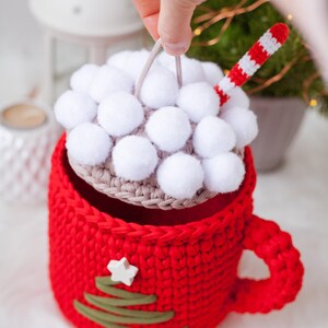Pu&ograve; includere: Una tazza rossa all'uncinetto con un design a forma di albero di Natale verde e una stella bianca. La tazza ha un coperchio rimovibile con pompon bianchi e una cannuccia a righe rosse e bianche.