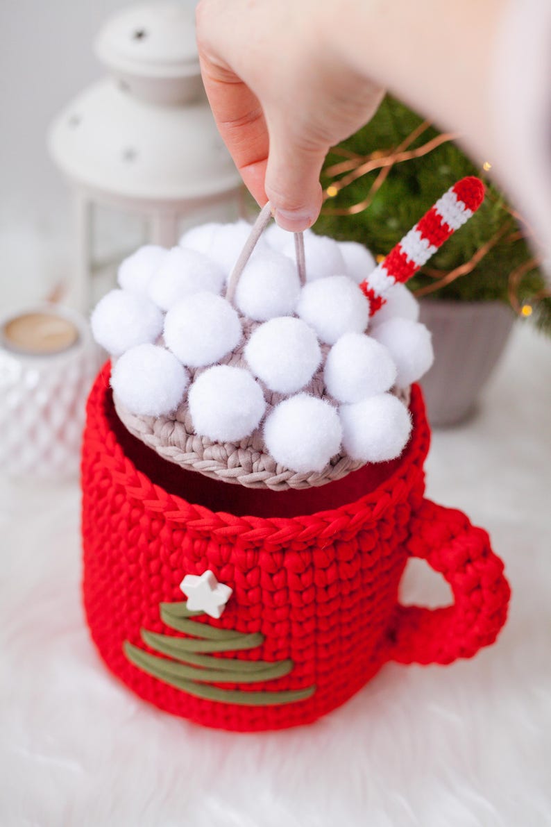 Pu&ograve; includere: Una tazza rossa all'uncinetto con un design a forma di albero di Natale verde e una stella bianca. La tazza ha un coperchio rimovibile con pompon bianchi e una cannuccia a righe rosse e bianche.