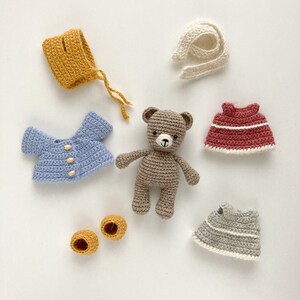 Op de afbeelding: Een bruine gehaakte teddybeer met een blauw gehaakt shirt, een gele gehaakte muts, een rode gehaakte jurk, een grijze gehaakte jurk, een witte gehaakte sjaal en twee gele gehaakte schoenen.