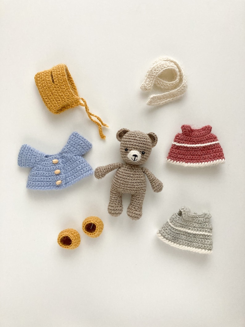 Op de afbeelding: Een bruine gehaakte teddybeer met een blauw gehaakt shirt, een gele gehaakte muts, een rode gehaakte jurk, een grijze gehaakte jurk, een witte gehaakte sjaal en twee gele gehaakte schoenen.