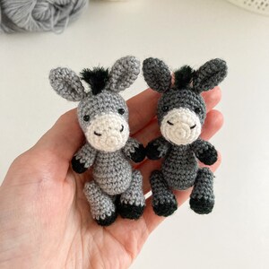 May include: Two gray crocheted donkey figurines with black hooves and black yarn for manes. The donkeys are sitting on a hand.