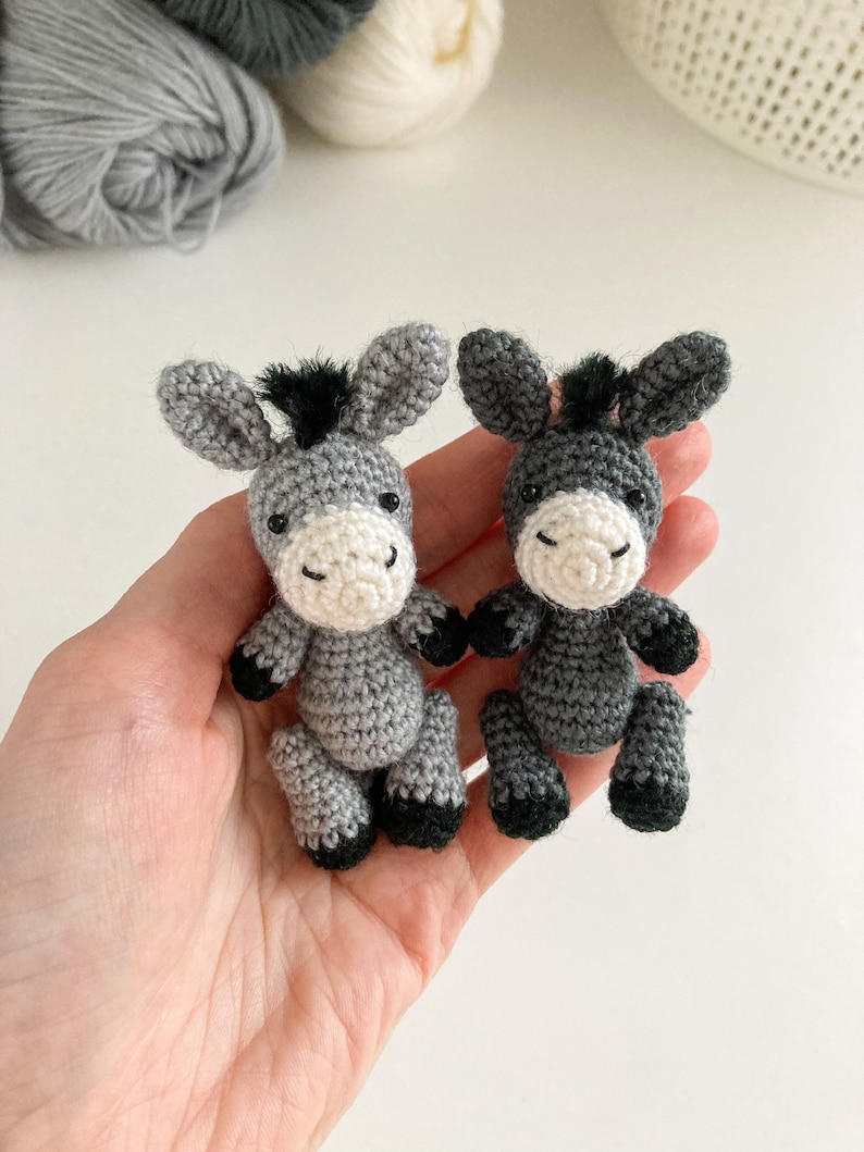 May include: Two gray crocheted donkey figurines with black hooves and black yarn for manes. The donkeys are sitting on a hand.