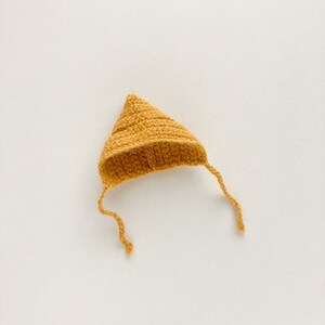 Op de afbeelding: Een kleine, gehaakte muts in een mosterdgele kleur. De muts heeft een puntige bovenkant en een lange stropdas die naar beneden hangt.