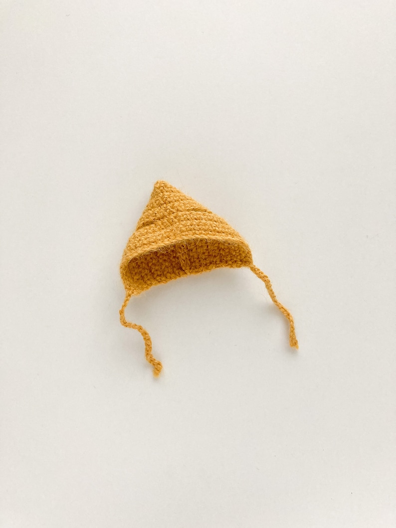 Op de afbeelding: Een kleine, gehaakte muts in een mosterdgele kleur. De muts heeft een puntige bovenkant en een lange stropdas die naar beneden hangt.