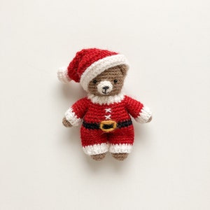 K&ouml;nnte beinhalten: Ein kleiner geh&auml;kelter Teddyb&auml;r, der eine rot-wei&szlig;e Weihnachtsm&uuml;tze und einen rot-wei&szlig;en Weihnachtsmann-Anzug mit einem schwarzen G&uuml;rtel tr&auml;gt.