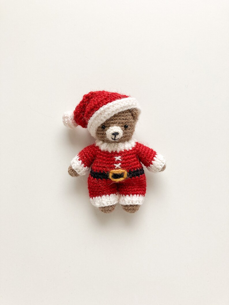 K&ouml;nnte beinhalten: Ein kleiner geh&auml;kelter Teddyb&auml;r, der eine rot-wei&szlig;e Weihnachtsm&uuml;tze und einen rot-wei&szlig;en Weihnachtsmann-Anzug mit einem schwarzen G&uuml;rtel tr&auml;gt.