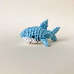 May include: A light blue crocheted shark toy with black eyes and a white belly. The shark is lying on its side with its tail and fins visible.