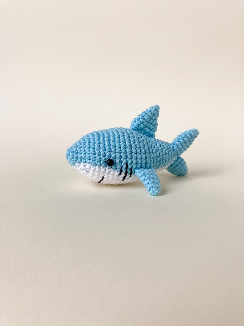 May include: A light blue crocheted shark toy with black eyes and a white belly. The shark is lying on its side with its tail and fins visible.