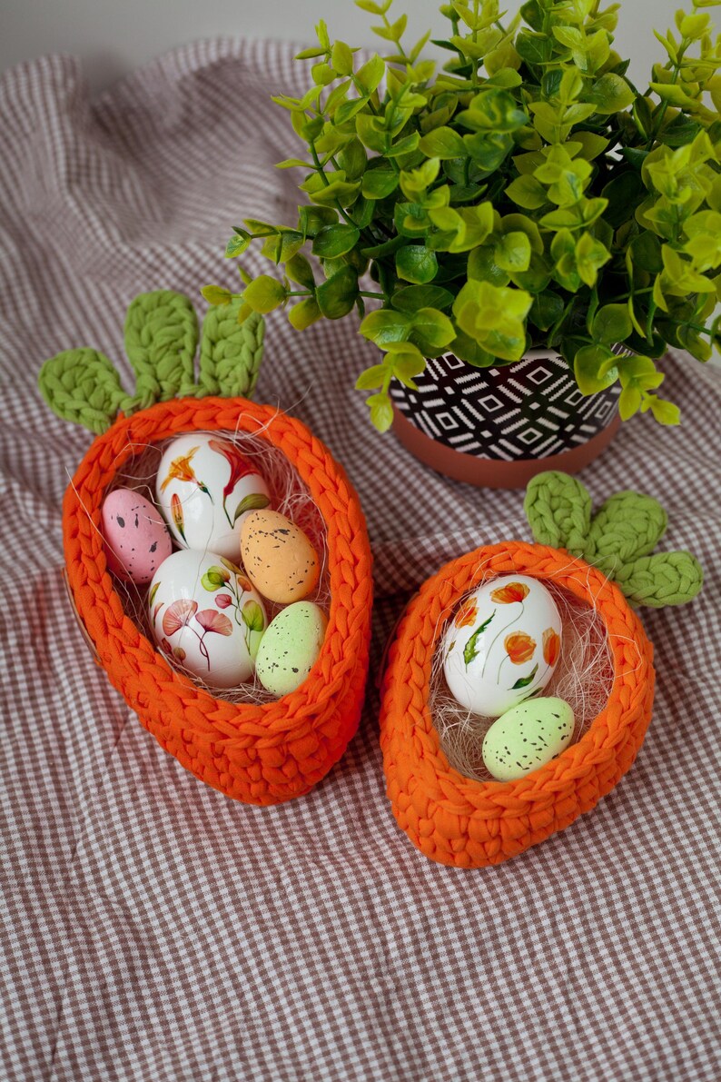 May include: Two orange crocheted carrot-shaped baskets filled with colorful Easter eggs. The baskets are on a brown and white checkered tablecloth.