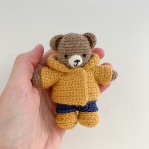 May include: A small brown crocheted teddy bear wearing a yellow crocheted jacket and blue crocheted pants. The bear is holding its hands in front of it.
