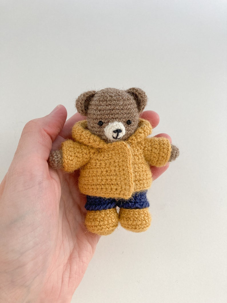 May include: A small brown crocheted teddy bear wearing a yellow crocheted jacket and blue crocheted pants. The bear is holding its hands in front of it.