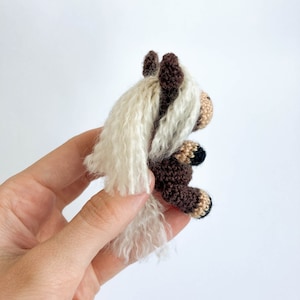 May include: A handmade crochet horse figurine. The horse has a brown body, a beige muzzle, and a white mane and tail. The horse's hooves are black and beige. The figurine is held against a white background.
