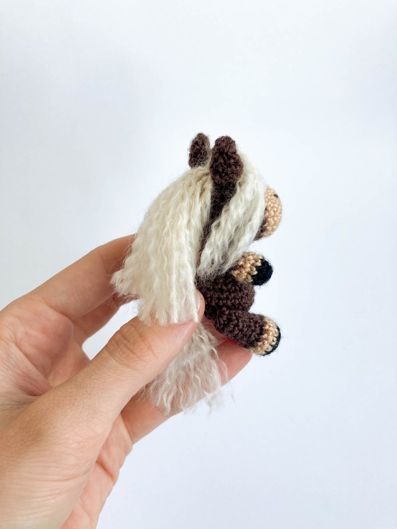 May include: A handmade crochet horse figurine. The horse has a brown body, a beige muzzle, and a white mane and tail. The horse's hooves are black and beige. The figurine is held against a white background.