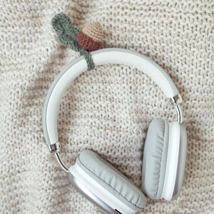 May include: White wireless headphones with gray earcups on a beige knitted blanket. A small crocheted acorn with a green leaf is attached to the headphones.