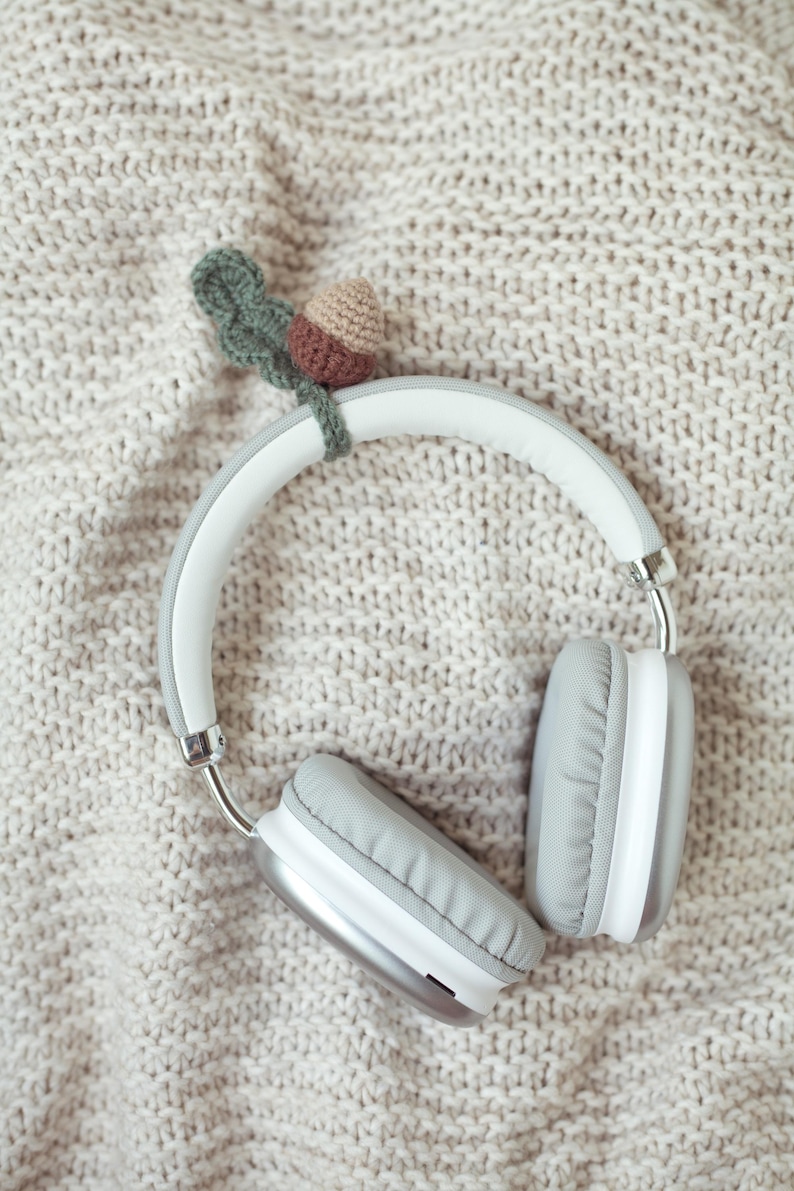 May include: White wireless headphones with gray earcups on a beige knitted blanket. A small crocheted acorn with a green leaf is attached to the headphones.