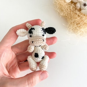 May include: A small crocheted cow toy with black and white spots. The cow is sitting in a hand with its head tilted up.