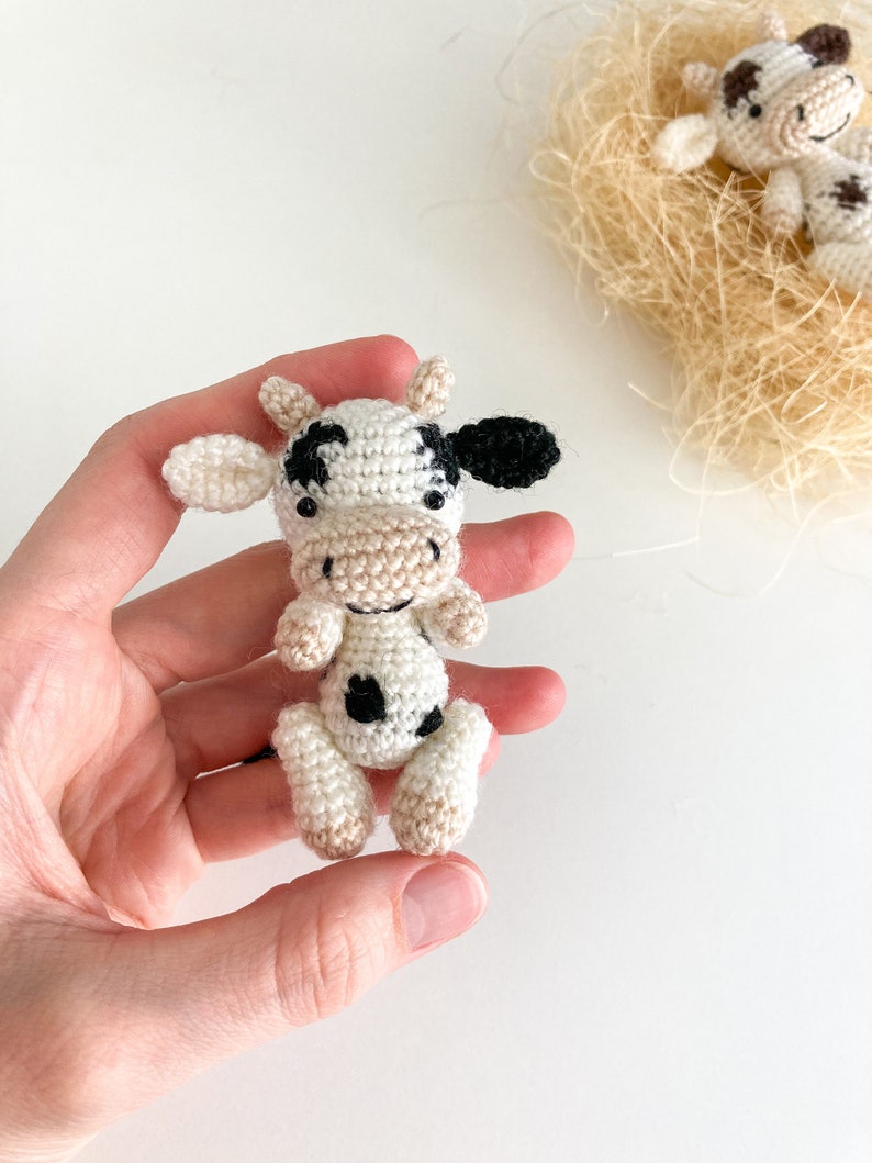 May include: A small crocheted cow toy with black and white spots. The cow is sitting in a hand with its head tilted up.