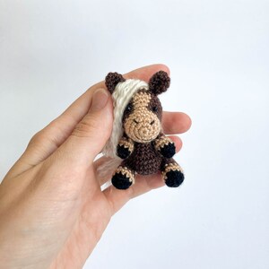 May include: A small, crocheted horse figurine. The horse has a brown body with a tan face and white mane. The hooves are black. The horse is sitting upright, with its legs tucked beneath it. The background is plain white.