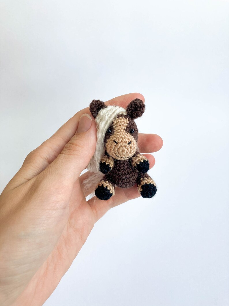 May include: A small, crocheted horse figurine. The horse has a brown body with a tan face and white mane. The hooves are black. The horse is sitting upright, with its legs tucked beneath it. The background is plain white.