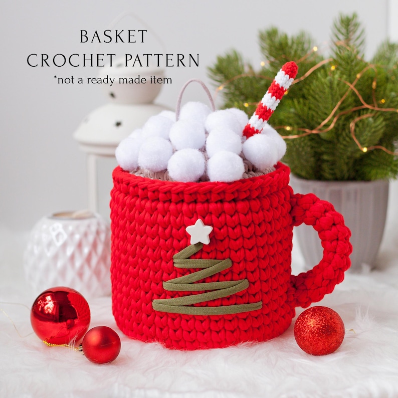 Pu&ograve; includere: Cestino rosso a forma di tazza lavorato a uncinetto con una stella bianca e un design a forma di albero verde. Il cestino &egrave; pieno di pompon bianchi e una cannuccia a righe rosse e bianche. Il cestino &egrave; appoggiato su una superficie bianca con ornamenti rossi.