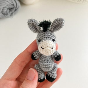 May include: A small gray crocheted donkey with black hooves and a black mane. The donkey is being held in a person's hand.