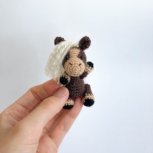 May include: A small, crocheted horse figurine with a brown body, tan face, and a white mane. The horse has black hooves and is waving its front leg. The toy is handmade and has a friendly, cartoonish appearance.