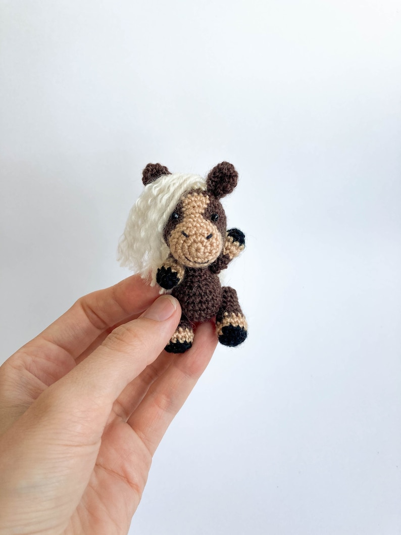May include: A small, crocheted horse figurine with a brown body, tan face, and a white mane. The horse has black hooves and is waving its front leg. The toy is handmade and has a friendly, cartoonish appearance.
