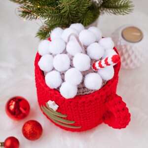 Pu&ograve; includere: Una tazza rossa all'uncinetto con un top a pompon bianco e una canna da zucchero che sporge. La tazza ha un design a forma di albero di Natale verde all'uncinetto sul lato. Ci sono ornamenti natalizi rossi su una superficie bianca e soffice.