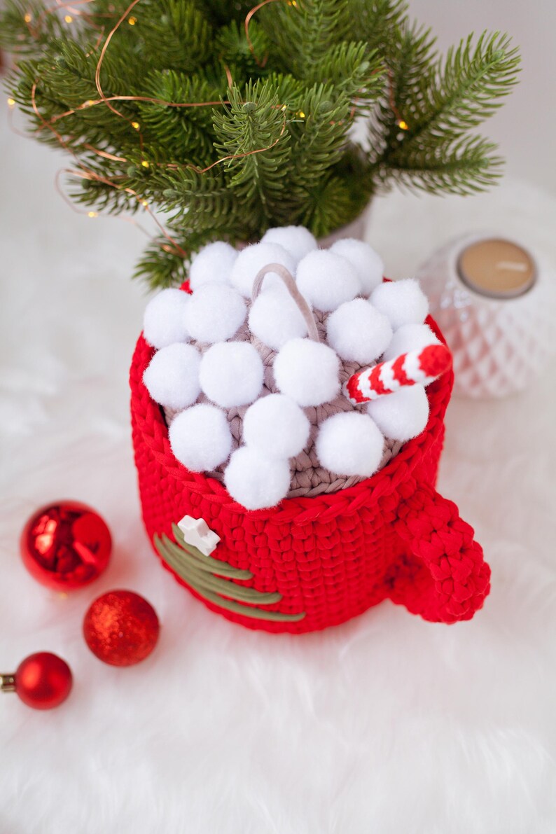 Pu&ograve; includere: Una tazza rossa all'uncinetto con un top a pompon bianco e una canna da zucchero che sporge. La tazza ha un design a forma di albero di Natale verde all'uncinetto sul lato. Ci sono ornamenti natalizi rossi su una superficie bianca e soffice.