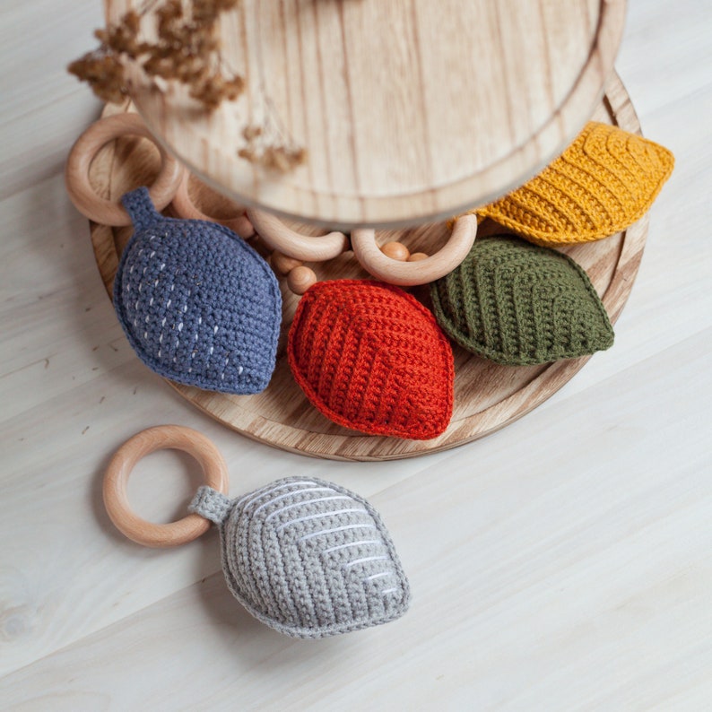 May include: Five crocheted leaf-shaped teethers in different colors, including gray, blue, red, green, and yellow. The teethers are arranged on a wooden tiered stand.
