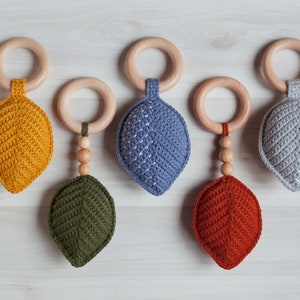 May include: Five crocheted leaf-shaped teethers with wooden rings and beads. The teethers are in shades of yellow, blue, green, red, and gray.