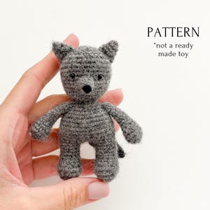 May include: A small, gray crocheted wolf toy with a black nose and small ears. The text "PATTERN *not a ready made toy" is visible in the upper right corner. The toy is held in a person's hand.