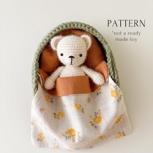 May include: A crocheted white teddy bear nestled in a green and orange bed with a floral patterned blanket. The text "PATTERN *not a ready made toy" is visible. The bear has black eyes and a black "T" shaped mouth.