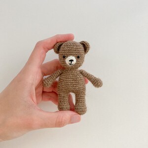 May include: A small brown crocheted teddy bear with black eyes and a white nose. The bear is being held in a hand.