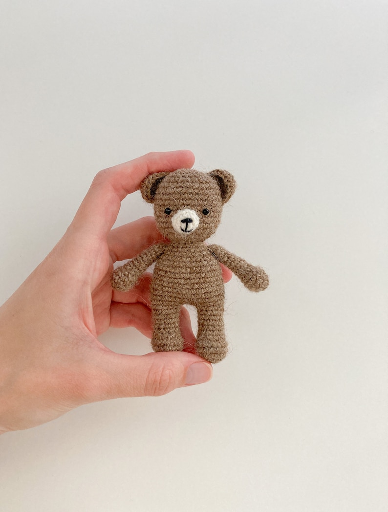 May include: A small brown crocheted teddy bear with black eyes and a white nose. The bear is being held in a hand.