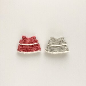 Op de afbeelding: Twee gehaakte poppenjurkjes, &eacute;&eacute;n in een koraalkleur en &eacute;&eacute;n in lichtgrijs. Beide jurkjes hebben een witte gehaakte rand.