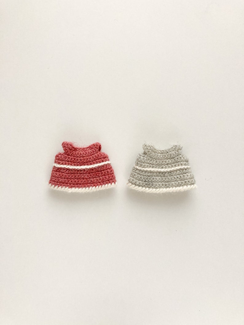 Op de afbeelding: Twee gehaakte poppenjurkjes, &eacute;&eacute;n in een koraalkleur en &eacute;&eacute;n in lichtgrijs. Beide jurkjes hebben een witte gehaakte rand.